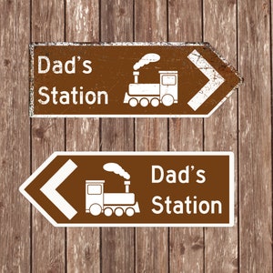 May include: Two brown wooden signs with white text and a white train illustration. The top sign reads "Dad's Station" with an arrow pointing to the right. The bottom sign reads "Dad's Station" with an arrow pointing to the left.