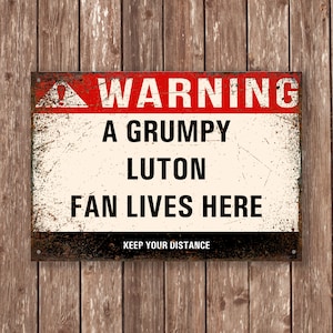 May include: A rectangular metal sign with a distressed finish. The sign has a red border at the top with the word "WARNING" in white. Below, the text reads "A GRUMPY LUTON FAN LIVES HERE" in black. The bottom of the sign says "KEEP YOUR DISTANCE".