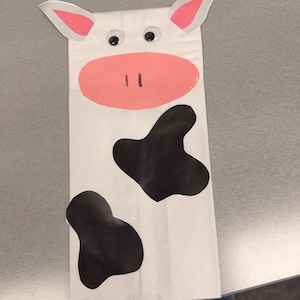 May include: A white paper bag craft shaped like a cow, featuring two googly eyes, a large pink oval snout with two vertical black nostrils, two pointed ears with pink centers, and two irregular black spots on the lower portion of the bag.