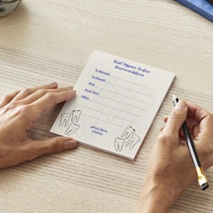 May include: A notepad with the text "Oral Hygiene Routine Recommendations" and tooth illustrations. The notepad is being held by a person's hands, with a pen in the other hand. The notepad has lines for writing.