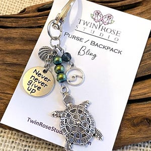 May include: Silver charm keychain with a turtle, seashell, and beads. The charm also has a silver disc with the words "Never never give up". The keychain is attached to a silver key ring. The keychain is on a white card with the text "Twin Rose Studio Purse/Backpack Bling".