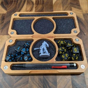 May include: A wooden box with a black felt interior and a compartment for dice and a miniature figure. The box has a pen holder and is designed for tabletop gaming.