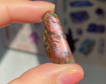 May include: A small, iridescent gemstone with brown, green, and pink tones. The stone is held between two fingers, highlighting its natural, rough texture. The background features blurred blue flowers and a colourful chart.