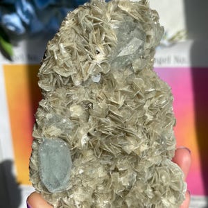 May include: A light-coloured, rough-textured mineral specimen with embedded blue-green crystals. The mineral has a layered appearance, with a cluster of small, flat crystals. The specimen is held in a hand, with a colourful background.