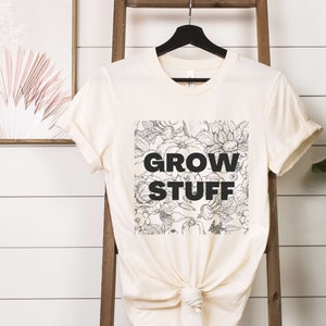 May include: A cream colored t-shirt with a black and white floral design and the text "GROW STUFF" printed on the front.