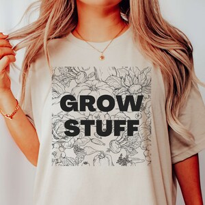 May include: A beige t-shirt with a black and white graphic of vegetables and flowers. The graphic includes the text "GROW STUFF".