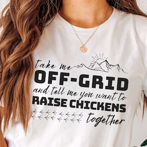 May include: White t-shirt with black text that reads "Take me off-grid and tell me you want to raise chickens together". The text is surrounded by a graphic of a mountain range and a sun.