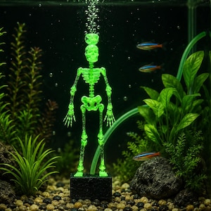 Op de afbeelding: Een neon-groen skelet aquariumornament staat op een zwarte rechthoekige voet in een aquarium. Het skelet is centraal geplaatst, omringd door groene waterplanten, donkere rotsen en kleine steentjes, met luchtbellen die vanaf de bovenkant opstijgen.