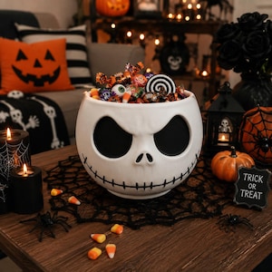 May include: A white ceramic bowl shaped like a skull with large black eye sockets and a stitched mouth, filled with assorted Halloween candies including candy corn, chocolate wrappers, and a black and white spiral lollipop.