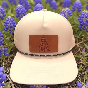 May include: A cream-colored trucker hat with a brown leather patch featuring a plant design. The hat has a black and white rope detail and a curved brim. The background is filled with vibrant blue flowers.
