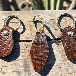 May include: Three brown leather keychains with silver and gold key rings. Each keychain has a unique shape and embossed pattern. The keychains are attached with metal rivets.