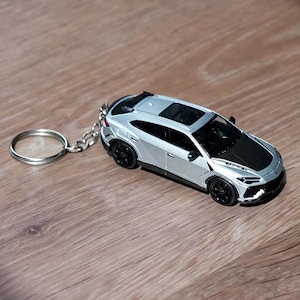 May include: A silver and black die-cast model car keychain. The miniature SUV has black wheels and a black hood. The keychain ring and chain are silver. The car is on a wooden surface.
