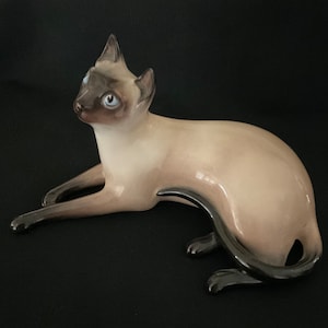 Op de afbeelding: Een porseleinen beeldje van een Siamese kat die ligt met zijn hoofd naar de zijkant gedraaid. De kat is lichtbruin met een zwarte staart en blauwe ogen.