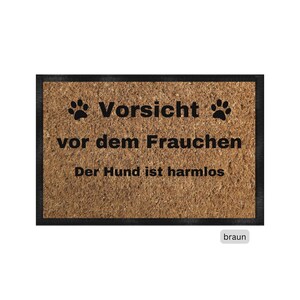 Può includere: Un tappetino da porta marrone con testo nero che recita "Vorsicht vor dem Frauchen Der Hund ist harmlos" con due impronte di zampa su entrambi i lati della parola "Vorsicht".