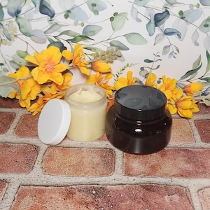 May include: Two jars of cream, one with a white lid and one with a black lid. The jar with the white lid is filled with a light yellow cream. The jar with the black lid is filled with a dark brown cream. The jars are sitting on a brick surface.