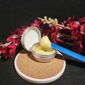 May include: A small silver tin with a yellow cream inside, being scooped out with a blue spatula. The tin is sitting on a cork coaster. The background is a black surface with a red flower arrangement.