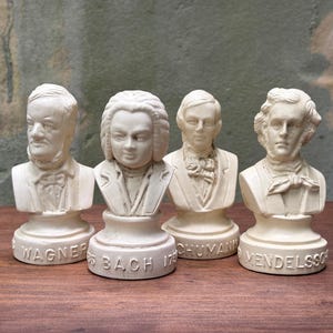May include: A set of four off-white, monochromatic busts of classical composers. Each figurine features a sculpted head and shoulders on a circular base. The names Wagner, Bach, Schumann, and Mendelssohn are embossed on the front of each base.