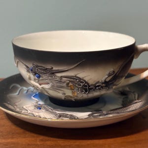 May include: A porcelain teacup and saucer set featuring a raised, three-dimensional dragon motif in shades of gray, black, and white. The dragon has bright blue eyes and is depicted wrapping around the cup and across the saucer surface.