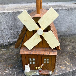 May include: A wooden windmill figurine featuring a brown structure with a textured roof, a central rotating blade assembly in cream, two small windows, a door, a white sack labeled Flour, and a small potted plant with yellow flowers at the base.