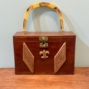 May include: A rectangular wooden box handbag with a dark brown finish, featuring a gold-tone metal latch, a central fleur-de-lis emblem, and two diamond-shaped carved floral panels. It has a curved, translucent amber-colored tortoiseshell handle.