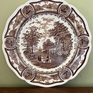 May include: A white and brown decorative plate with a scalloped edge. The plate features a detailed landscape scene with figures, trees, and buildings. The border includes decorative elements. The plate is likely vintage or antique.