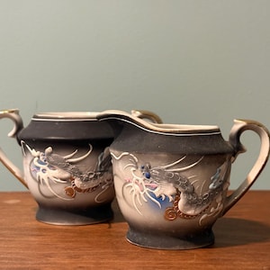 May include: A ceramic creamer and sugar bowl set featuring a matte black and white gradient finish. Each piece is decorated with a raised, three-dimensional dragon motif in shades of gray, blue, and gold, accented with gold-trimmed handles.