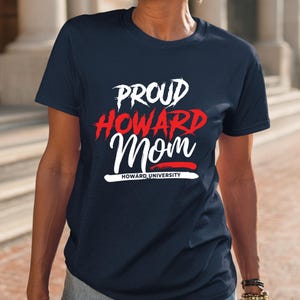 May include: Navy blue t-shirt with the words "PROUD HOWARD Mom" in white and red lettering. The text "HOWARD UNIVERSITY" is printed in white below the word "Mom". The shirt is a crew neck style.
