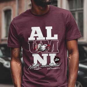 May include: Maroon t-shirt with white text that reads "ALUMNI" stacked vertically. The "U" features a graphic of an eagle and a building. Below the text is "North Carolina Central University" and "EST 1910".