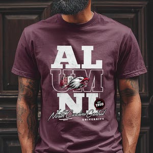 May include: Maroon t-shirt with the word "ALUMNI" in white block letters. The shirt features a graphic of an eagle and the text "North Carolina Central University" in a script font. The year "1910" is also printed on the shirt.