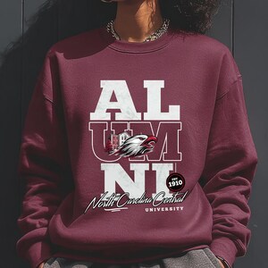 May include: Burgundy sweatshirt with the word "ALUMNI" in white block letters. A graphic of an eagle and a building is centered within the letters. Below the text is "North Carolina Central University" and "EST 1910" in a circular design.