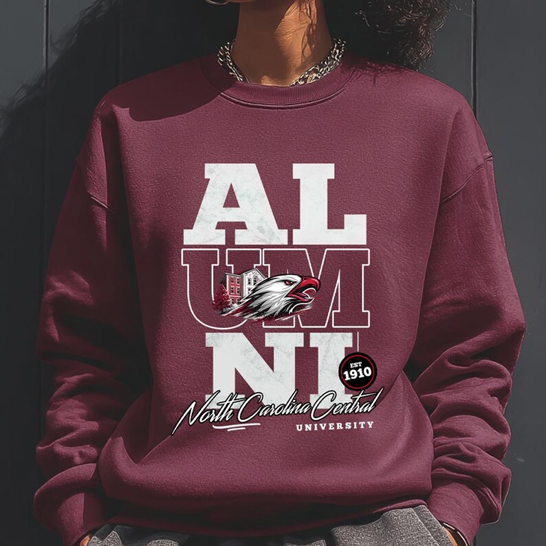 May include: Burgundy sweatshirt with the word "ALUMNI" in white block letters. A graphic of an eagle and a building is centered within the letters. Below the text is "North Carolina Central University" and "EST 1910" in a circular design.