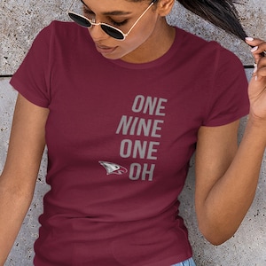 May include: Burgundy t-shirt with the words "ONE NINE ONE OH" in silver lettering. A stylized bird logo is below the text. The shirt is a crew neck and appears to be made of cotton.