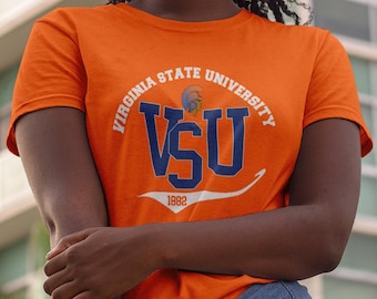 Può includere: T-shirt arancione con il logo della Virginia State University. Il design include le lettere "VSU" in blu navy, il nome dell'università in bianco e l'anno "1882". La maglietta è a maniche corte.