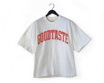 May include: White t-shirt with the word "GOODTASTE" printed in red, arched across the chest. The shirt is hanging on a black hanger against a white background. The t-shirt has short sleeves and a crew neck.