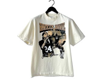 May include: Cream-colored t-shirt with a high neckline, featuring a graphic of a person wearing a jersey with the number 24. The shirt has the text "BREEZY BOWL" in a gold font. The graphic also includes the signature "Chris Brown".
