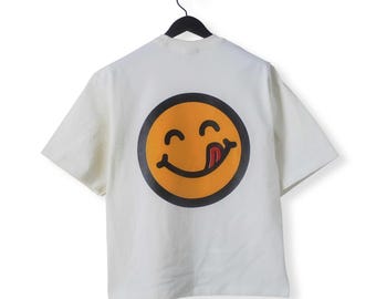 May include: Off-white short-sleeve t-shirt with a large, orange smiley face graphic on the back. The smiley face has a black outline and a tongue sticking out. The shirt is hanging on a black hanger.