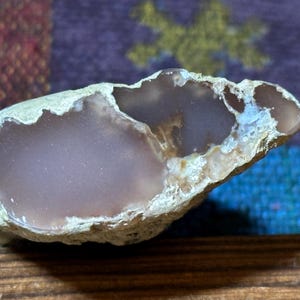 May include: A natural, irregularly shaped stone with a smooth, light purple interior and a rough, off-white exterior. The stone has a unique, organic form, with a hollowed-out section. The stone is displayed on a wooden surface.