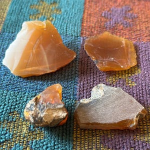 May include: Four raw, uncut gemstones in shades of orange and tan are displayed on a colorful patterned fabric. The stones vary in size and texture, with some appearing translucent and others opaque.