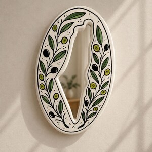 May include: An oval-shaped wall mirror with a white frame featuring a hand-painted design of green olive branches, leaves, and black olives. The mirror glass is shaped like the map of Israel, creating a unique cutout effect within the oval frame.