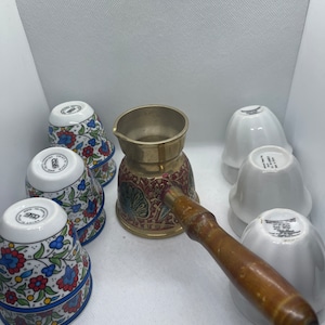 May include: A set of six traditional Turkish coffee cups with floral patterns and a brass cezve with a wooden handle. The cups are stacked in pairs and the cezve is in the center of the image.