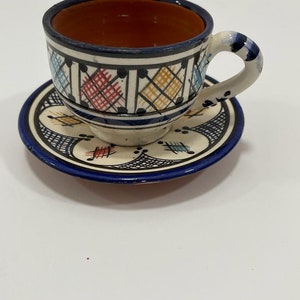 Może przedstawiać: Biały ceramiczny kubek do herbaty i spodek z niebieskimi, czerwonymi i żółtymi wzorami geometrycznymi. Kubek ma brązowe wnętrze.