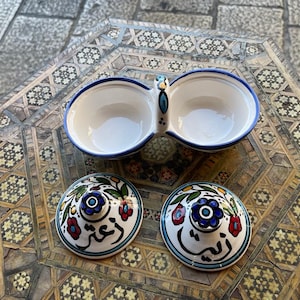 May include: A white ceramic double dish with blue trim and floral designs. The dish has two separate bowls and a handle in the middle. The dish is sitting on a colorful mosaic surface. The dish has two lids with floral designs and Arabic script.