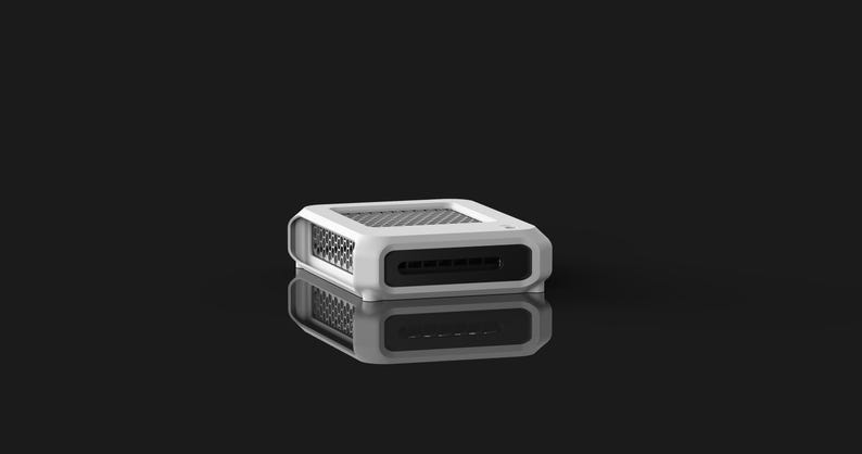 Op de afbeelding: Een wit en zwart rechthoekig elektronisch apparaat met een mesh-bovenkant en een reeks sleuven aan de voorkant. Het apparaat rust op een reflecterend oppervlak, waardoor een spiegeleffect ontstaat tegen een zwarte achtergrond.