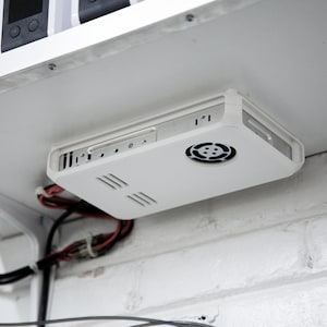 Op de afbeelding: Een wit elektronisch apparaat met een ventilator en ventilatiesleuven is onder een witte plank gemonteerd. Draden en kabels zijn zichtbaar. Het apparaat is bevestigd aan een witte bakstenen muur.