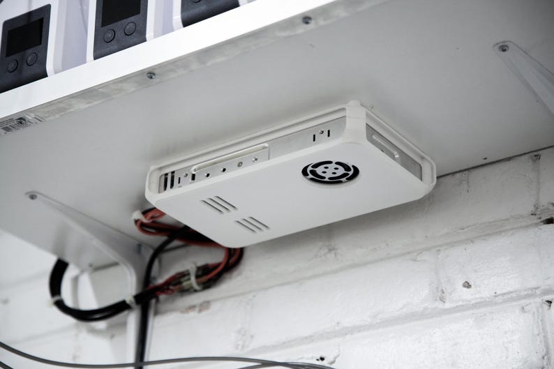 Op de afbeelding: Een wit elektronisch apparaat met een ventilator en ventilatiesleuven is onder een witte plank gemonteerd. Draden en kabels zijn zichtbaar. Het apparaat is bevestigd aan een witte bakstenen muur.