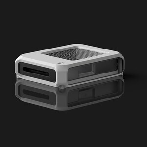 Op de afbeelding: Een wit en zwart rechthoekig elektronisch apparaat met een mesh-bovenkant en een reflecterend oppervlak eronder. Het apparaat heeft een sleuf aan de voorkant en een doorzichtig zijpaneel. De achtergrond is zwart.