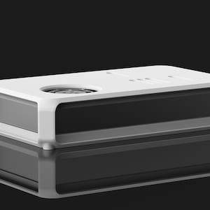 May include: A rectangular, modern-style device with a white top and a gray base. The top features a circular element and several small, rectangular indentations. The device is reflected on a glossy black surface, creating a symmetrical image.