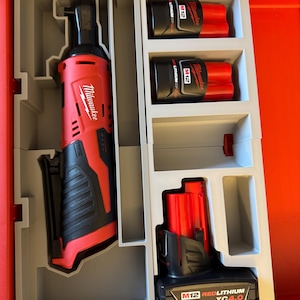 Puede incluir: Una llave de impacto inalámbrica Milwaukee roja y negra con una batería de 12V y una batería de iones de litio roja. Las herramientas están en una caja de herramientas gris y roja.