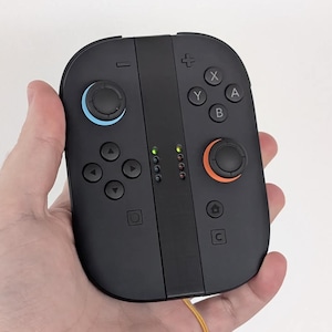 Pode incluir: Controlador de jogos portátil preto com joysticks azuis e laranja, botões direcionais e botões de ação. O controlador tem luzes indicadoras e é segurado em uma mão.