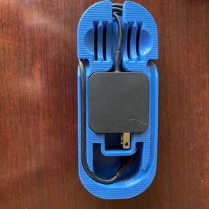 May include: A blue 3D-printed cable organizer with a black power adapter and black cables. The organizer has a rectangular shape with slots and curves to hold the cables and adapter. The item is on a wooden surface.